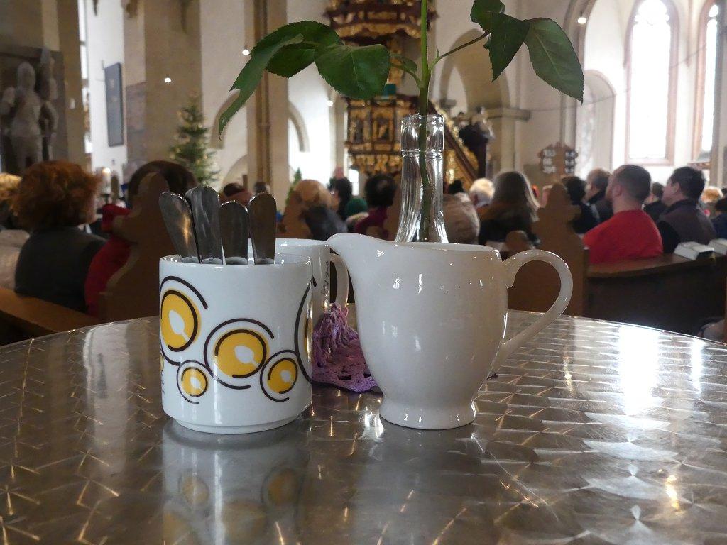 Eine Tasse mit Vesperkirche-Logo und ein Milchkännchen stehen auf einem Stehtisch im Vordergrund. Dahinter ist die Gottesdienstgemeinde zu erkennen.